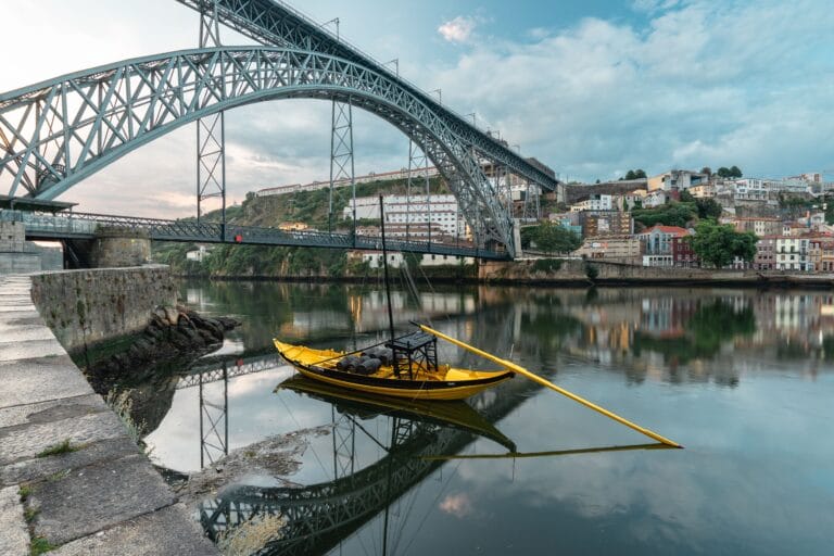 Wide view of a large steel arch bridge crossing a calm river with a yellow boat in the foreground and a hillside town along the shore
