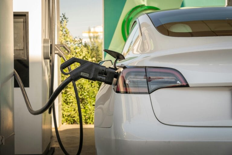 White electric car connected to a charging station with the plug inserted into the rear left side near the taillight