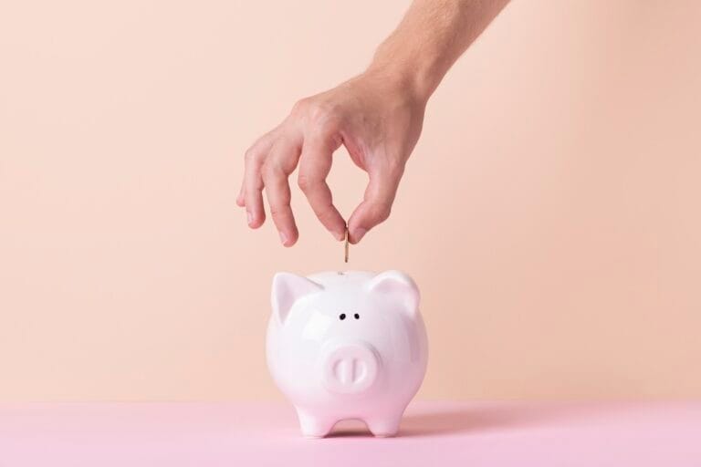 Hand placing a coin into a white pink piggy bank on a pale pink background
