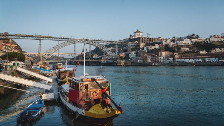 Colorful boats docked along a calm river with a large metal double deck arch bridge and hillside city in the background Porto