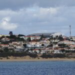 Coastal hillside town with white and pastel houses along a sandy beach under a cloudy sky and blue sea in foreground
