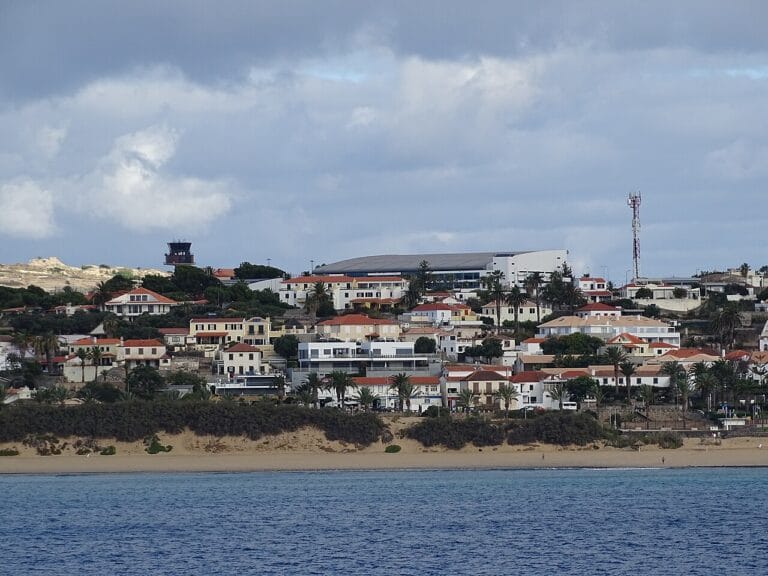 Coastal hillside town with white and pastel houses along a sandy beach under a cloudy sky and blue sea in foreground