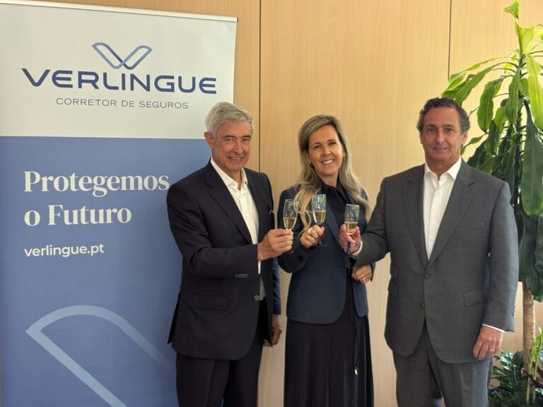 Three suited professionals toast with champagne in front of a Verlingue insurance banner that reads Protegemos o Futuro
