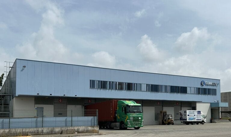Industrial warehouse with a light blue metal exterior loading bays and parked trucks under a cloudy sky