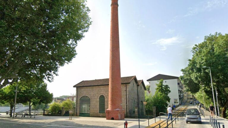 Tall red brick chimney beside a stone building in a sunny town square with trees and pedestrians nearby