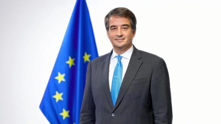 Man in a dark gray suit and blue tie stands beside a European Union flag against a white backdrop