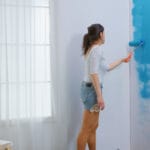Woman painting a white wall blue with a roller ladder and paint bucket nearby in a bright room with sheer curtains