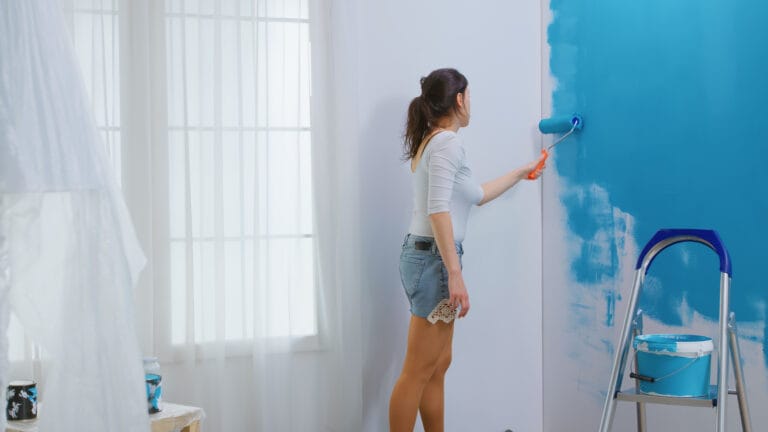 Woman painting a white wall blue with a roller ladder and paint bucket nearby in a bright room with sheer curtains