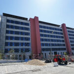 Construction site in front of a modern multi story building with pink columns red machinery and a gravel pile in the foreground