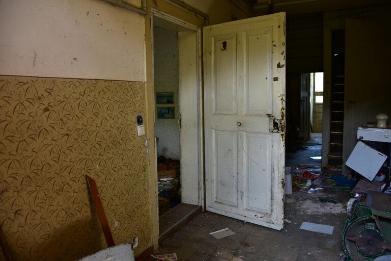 Abandoned interior with peeling wallpaper an open white door and clutter on the floor in dim light