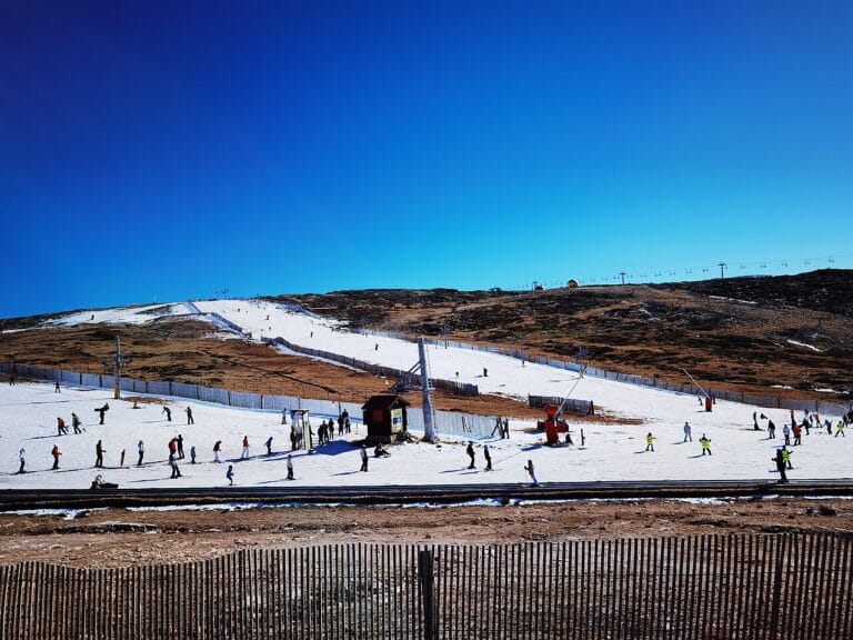 Crowded ski slope on a sunny day with a wooden fence in the foreground and a clear blue sky above the hill behind the slope under wintersnow