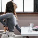 Woman in a gray sweater stands with hands on hips beside a laptop at a bright office desk stretching or leaning back in a white chair