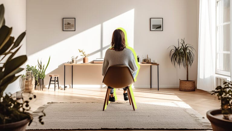 Person seated at a desk in a sunlit living room viewed from behind plants on the floor and on the desk
