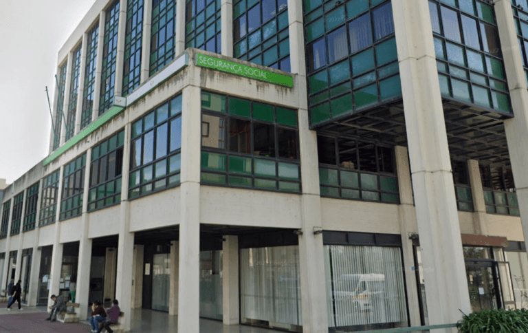 Exterior view of a government office building with green tinted windows and a Segurança Social sign people sit outside along the walkway