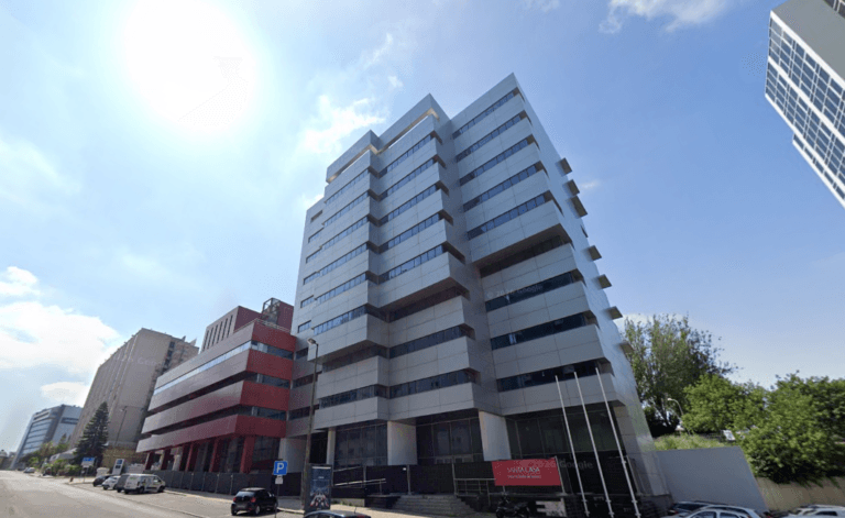 Tall modern office building with gray panels and glass windows on a sunny city street neighboring red bricktorso building and parked cars nearby