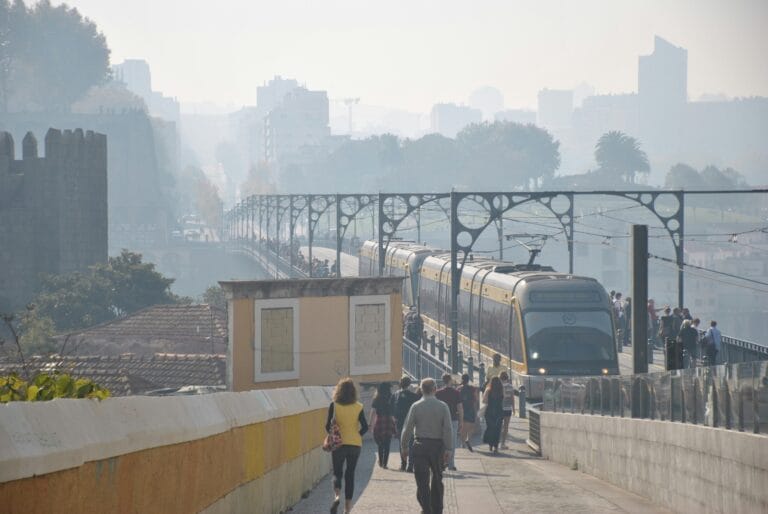 People walk along a barrier lined walkway beside an elevated metro train in a hazy urban landscape