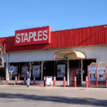 Staples storefront exterior with a red sign glass doors and a palm tree on the left under a clear blue sky