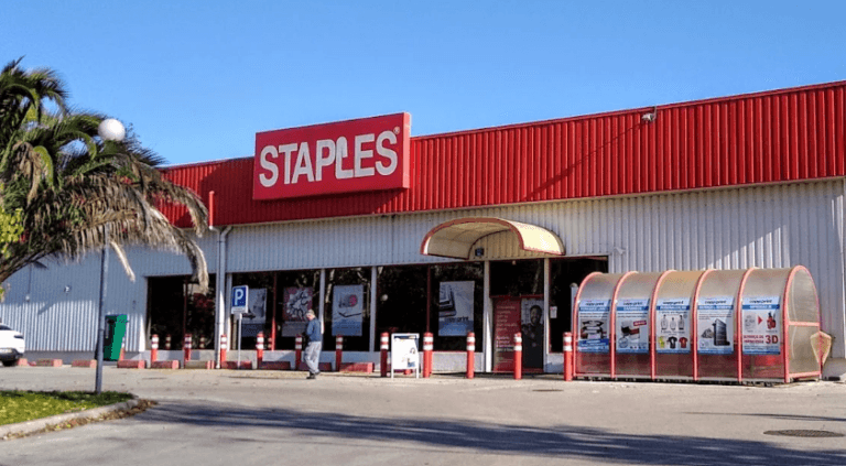 Staples storefront exterior with a red sign glass doors and a palm tree on the left under a clear blue sky