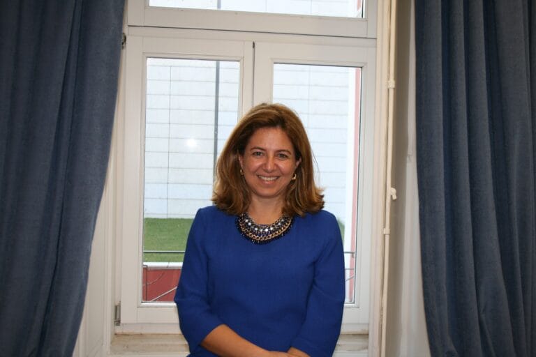 Smiling woman in a blue dress with a jeweled necklace standing by a window with dark curtains behind her