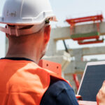 Construction worker in an orange safety vest and white helmet reviews a tablet checklist on a building site