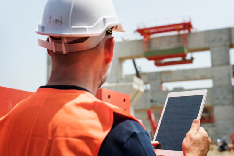 Construction worker in an orange safety vest and white helmet reviews a tablet checklist on a building site
