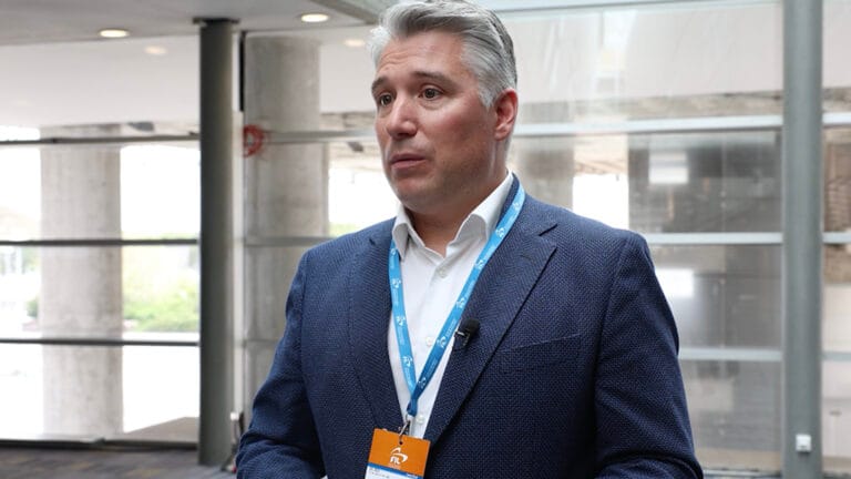Man in a blue blazer with white shirt wearing a conference lanyard and microphone speaking in a modern atrium