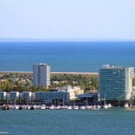 Coastal cityscape with a marina of boats in the foreground and modern buildings along the shore against a blue sea and sky horizon