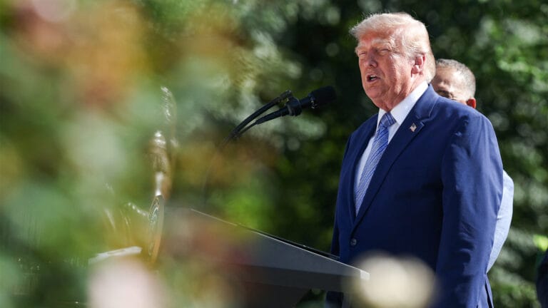 Man in a blue suit speaking into a microphone at an outdoor podium with greenery in the background