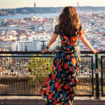 Woman in a bright floral dress stands at a railing overlooking a city with red tiled roofs river and distant bridge