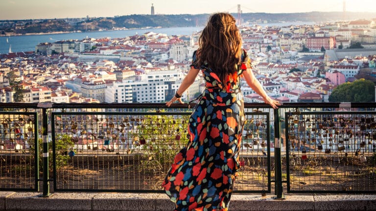 Woman in a bright floral dress stands at a railing overlooking a city with red tiled roofs river and distant bridge