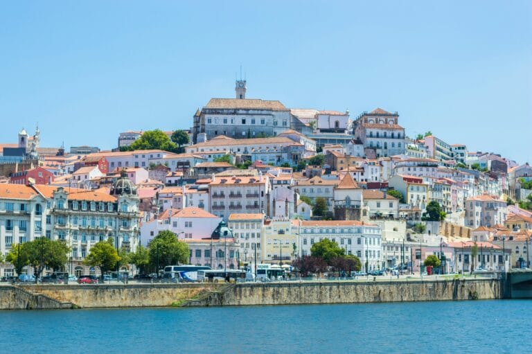 Hillside cityscape with pastel hued buildings and red tiled roofs along a river under a clear blue sky centered by a clock tower