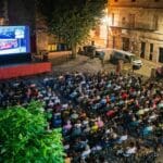 Open air nighttime cinema in a town square a large screen shows a red bus scene while a crowd sits in rows under string lights