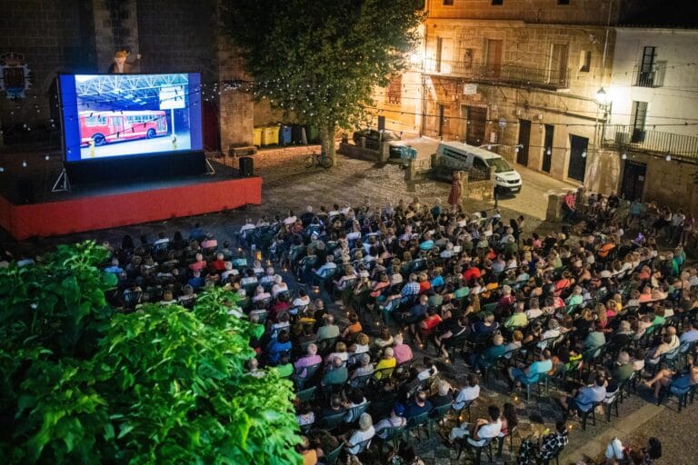 Open air nighttime cinema in a town square a large screen shows a red bus scene while a crowd sits in rows under string lights