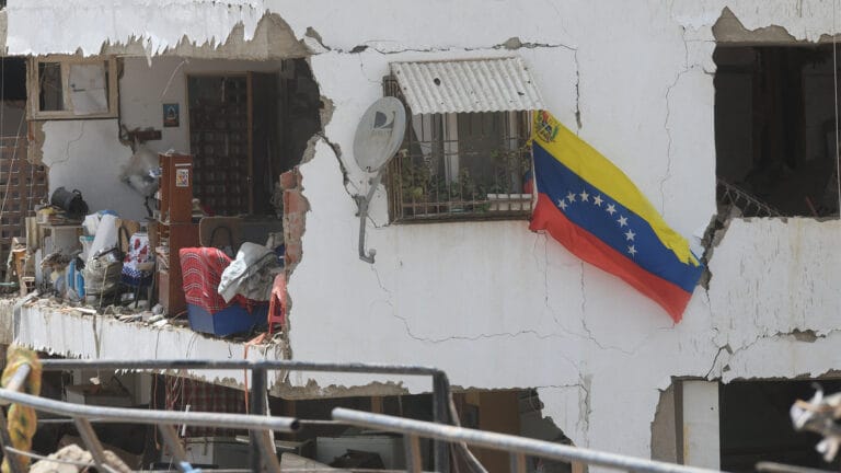 Ruined white building with cracked walls and a Venezuelan flag hanging from a windowrailing debris and household items scattered outside the rooms