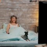 Woman resting in bed with light blue sheets smiling as a small dog sits beside her a TV screen appears in the foreground on the right