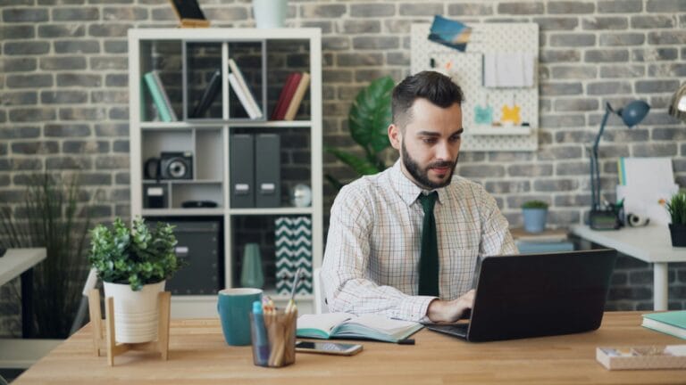 Man in a checkered shirt and green tie works on a laptop at a wooden desk in a modern brick office setting with plants in the foreground
