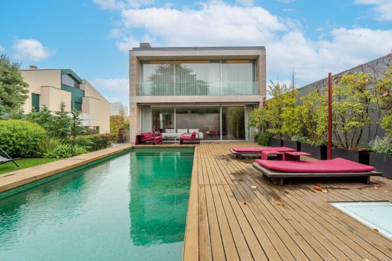 Modern twostory glass walled house with a wooden deck turquoise pool and red outdoor lounge chairs amid greenery