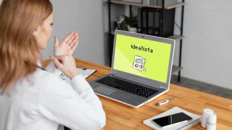 Person at a desk gestures toward a laptop displaying a lime green screen with the word idealista and a magnifying glass over papers logo shown