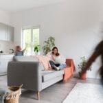 Woman lounging on a gray sofa with a laptop in a bright living room with plants and a pink throw blanket blurred children move in the background