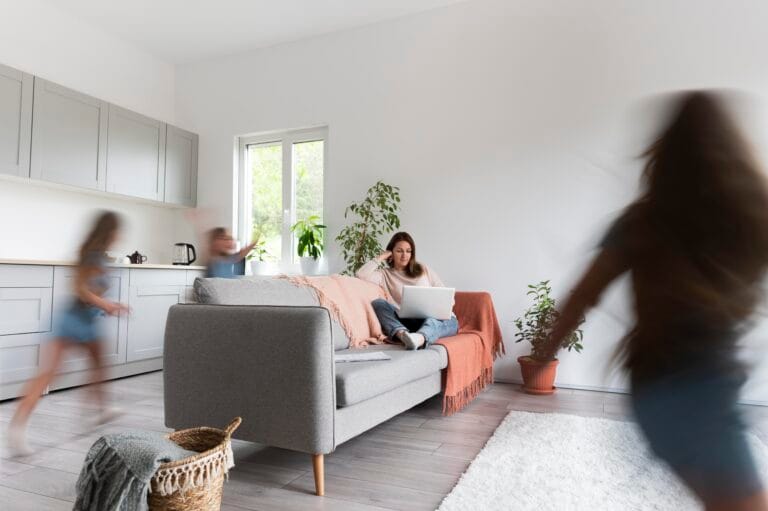 Woman lounging on a gray sofa with a laptop in a bright living room with plants and a pink throw blanket blurred children move in the background