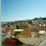 Panoramic view of a coastal town with terracotta rooftops white and pastel buildings and green hills under a clear blue sky seen from a window on the left The sea is visible to the far left