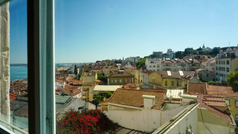 Panoramic view of a coastal town with terracotta rooftops white and pastel buildings and green hills under a clear blue sky seen from a window on the left The sea is visible to the far left