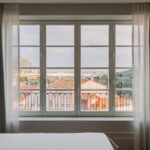 Bedroom with a large window and sheer curtains showing red rooftops and a distant bridge over water