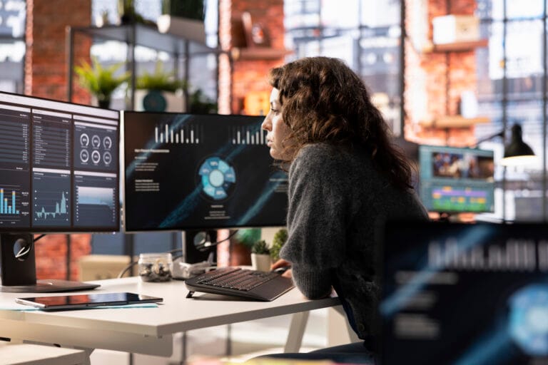 Person seated at a desk in an office looking at multiple monitors displaying charts and graphs